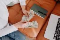 Seorang wanita tengah menghitung uang tunai di meja kerja yang penuh dengan perlengkapan bisnis kecil, menunjukkan semangat dan ketekunan dalam mengelola keuangan usaha dari awal. Gambar ini merepresentasikan fase awal pengumpulan modal dan pengelolaan finansial yang cermat bagi para perintis bisnis.
