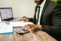 Seorang pemilik usaha mikro tengah fokus memeriksa laporan keuangan di laptopnya, menunjukkan dedikasi dalam mengelola aspek finansial demi keberlanjutan bisnisnya. Latar belakang tumpukan produk usaha mengindikasikan aktivitas bisnis yang sedang berjalan.