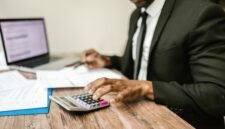 Seorang pemilik usaha mikro tengah fokus memeriksa laporan keuangan di laptopnya, menunjukkan dedikasi dalam mengelola aspek finansial demi keberlanjutan bisnisnya. Latar belakang tumpukan produk usaha mengindikasikan aktivitas bisnis yang sedang berjalan.
