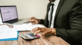 Seorang pemilik usaha mikro tengah fokus memeriksa laporan keuangan di laptopnya, menunjukkan dedikasi dalam mengelola aspek finansial demi keberlanjutan bisnisnya. Latar belakang tumpukan produk usaha mengindikasikan aktivitas bisnis yang sedang berjalan.