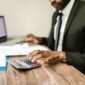Seorang pemilik usaha mikro tengah fokus memeriksa laporan keuangan di laptopnya, menunjukkan dedikasi dalam mengelola aspek finansial demi keberlanjutan bisnisnya. Latar belakang tumpukan produk usaha mengindikasikan aktivitas bisnis yang sedang berjalan.
