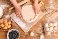 Seorang wanita sedang bekerja di meja dapur yang tertata rapi, dikelilingi oleh peralatan memasak dan bahan kerajinan, menggambarkan suasana produktif di rumah. Gambar ini merepresentasikan berbagai potensi usaha yang dapat dikembangkan dari rumah sendiri.