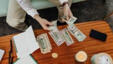 Seorang wanita tengah menghitung tumpukan koin yang dikumpulkan dalam toples, melambangkan upaya menabung secara konsisten untuk modal awal sebuah usaha. Gambar ini merefleksikan semangat wirausaha dalam mengelola keuangan pribadi.