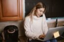Seorang wanita muda tengah serius bekerja di sudut meja rumahnya, dikelilingi perangkat digital dan produk-produk kreatif hasil karyanya. Foto ini merepresentasikan semangat wirausaha rumahan yang memanfaatkan teknologi untuk meraih kesuksesan.