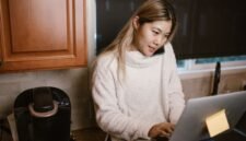 Seorang wanita muda tengah serius bekerja di sudut meja rumahnya, dikelilingi perangkat digital dan produk-produk kreatif hasil karyanya. Foto ini merepresentasikan semangat wirausaha rumahan yang memanfaatkan teknologi untuk meraih kesuksesan.