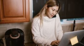 Seorang wanita muda tengah serius bekerja di sudut meja rumahnya, dikelilingi perangkat digital dan produk-produk kreatif hasil karyanya. Foto ini merepresentasikan semangat wirausaha rumahan yang memanfaatkan teknologi untuk meraih kesuksesan.